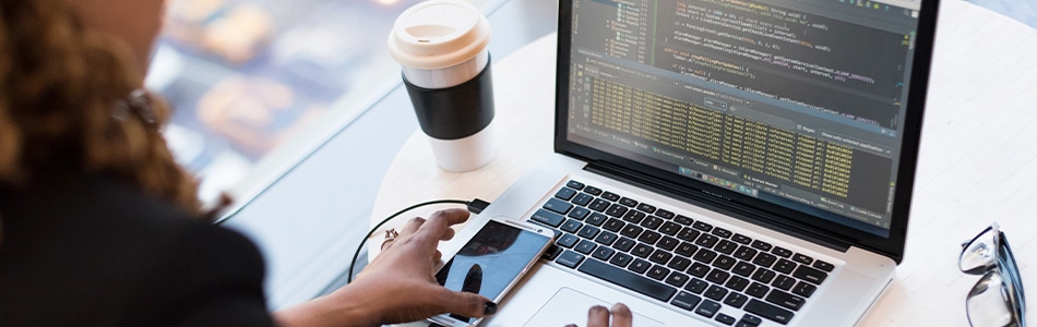 Imagem de um laptop preto e prata na mesa redonda de madeira marrom. O laptop está aberto e a tela está mostrando um código de programação. A mesa está também com um mouse, um teclado e um copo de café.