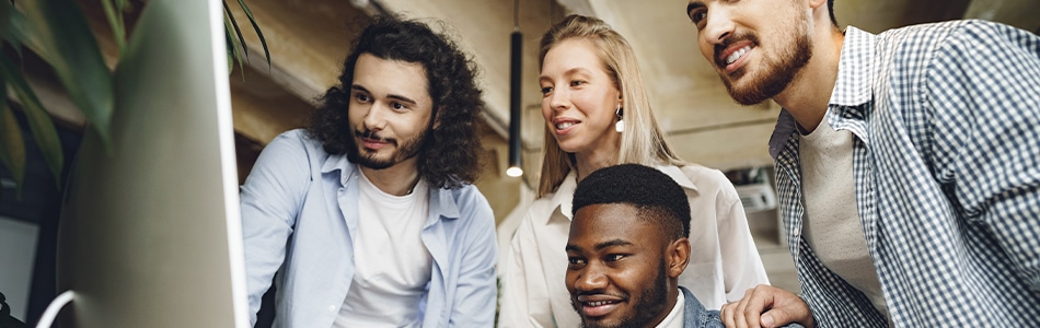Imagem de quatro colegas multiétnicos olhando para uma tela de computador em um escritório. Os colegas são homens e mulheres de diferentes idades e etnias. Eles estão vestidos de forma profissional e parecem estar concentrados no trabalho.