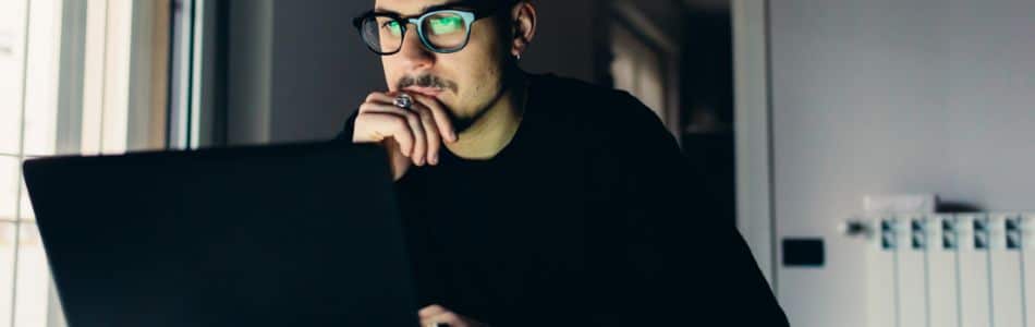Descrição de Imagem: Um homem de óculos e camisa preta olhando para o seu laptop.