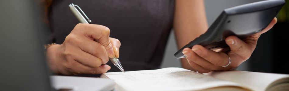Descrição de Imagem: Uma mão escrevendo em um papel e a outra segurando uma calculadora.