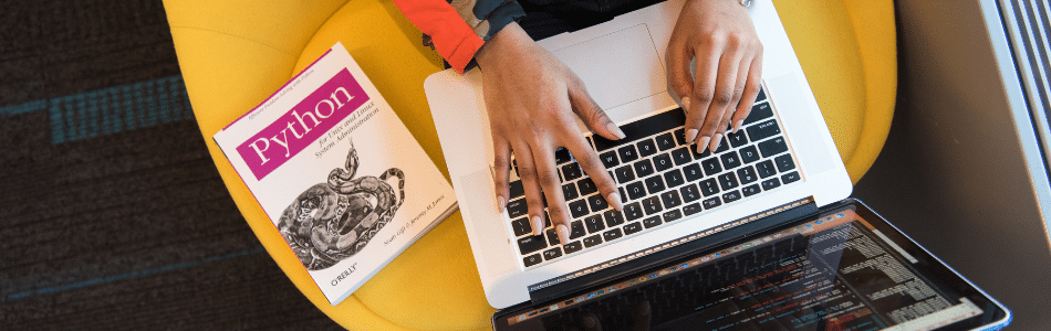 Descrição de Imagem: Uma pessoa trabalhando em um laptop com um código visível na tela, ao lado de um livro intitulado "python". apenas as mãos da pessoa e a capa do livro são visíveis.