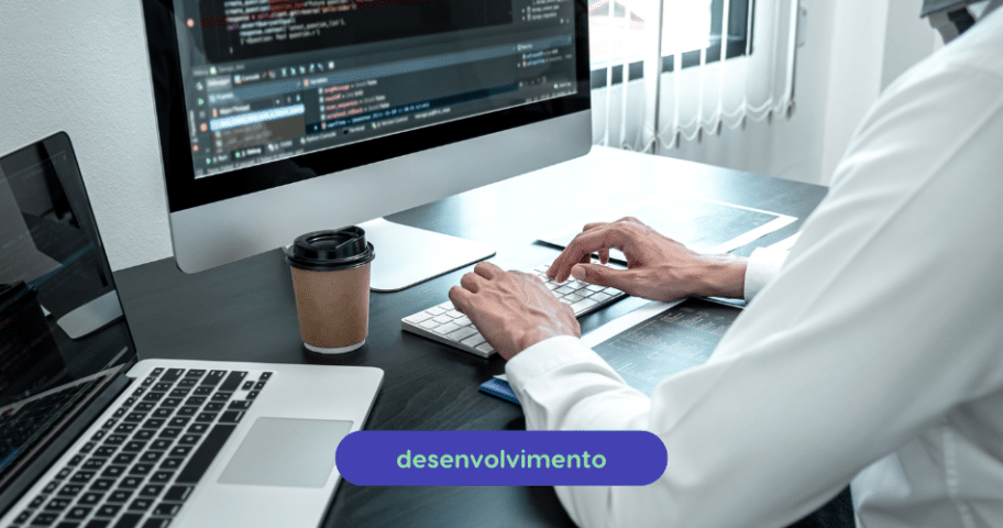 Uma pessoa está digitando em um teclado na frente de um computador desktop exibindo linhas de código. Há um laptop, uma xícara de café e um smartphone na mesa. A palavra “desenvolvimento” aparece em forma oval azul na parte inferior da imagem, enfatizando Qualidade de Software.