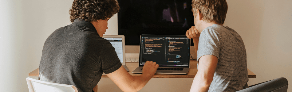 Descrição de Imagem: mesa trabalhando em um laptop exibindo código. Uma pessoa está apontando para a tela, discutindo APIs, enquanto a outra observa atentamente. Há outro laptop aberto ao lado deles na mesa, que também mostra algum tipo de texto ou código, destacando diferenças em serviços web.