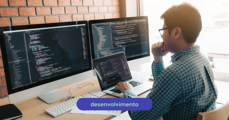 Descrição de Imagem: Um homem de óculos e camisa xadrez está sentado em uma mesa, olhando para várias telas de computador cheias de códigos e exercícios de Python. Ele também está usando um laptop.