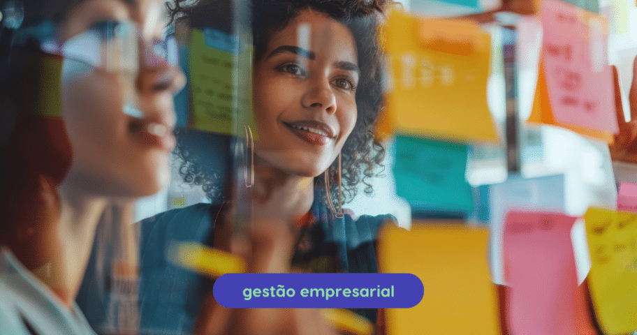 Descrição de Imagem: Uma pessoa com cabelo cacheado está olhando para uma parede de vidro coberta com post-its coloridos, parecendo pensativa e focada, pois seu reflexo é visível no vidro. O texto "gestão empresarial" e "Metodologias Ágeis" estão escritos em caixas de texto azuis na parte inferior da imagem.