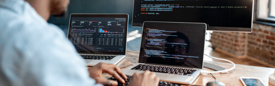 Descrição de Imagem: Uma pessoa trabalhando em dois laptops e um monitor, digitando no teclado de um laptop. Os laptops exibem análises de dados e linhas de código, provavelmente utilizando ferramentas de produtividade para JavaScript, enquanto o monitor mostra mais código. O cenário parece ser um escritório ou área de trabalho com janelas em segundo plano.