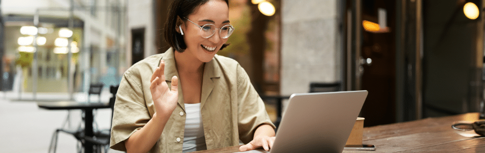 Descrição de Imagem: Uma mulher com óculos e fones de ouvido está sentada em uma mesa ao ar livre, sorrindo e acenando para a tela do seu laptop, provavelmente discutindo CS e CX em uma videochamada. Ela está usando uma camisa bege e o cenário parece ser um café ou outro espaço público.