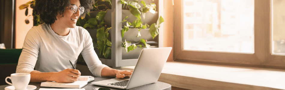 Descrição de Imagem: Uma pessoa com cabelo cacheado e óculos está sentada à mesa, sorrindo enquanto aprimora suas habilidades técnicas em um laptop. Tomando notas em um caderno com uma caneta, ela está cercada por uma planta em vaso e uma xícara. A luz do sol entra por uma grande janela, iluminando seu espaço de trabalho focado.