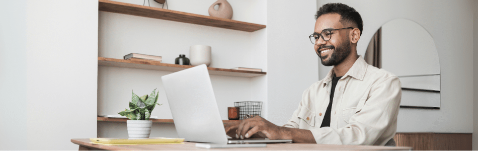 Descrição de Imagem: Um homem de óculos está sentado em uma mesa de madeira, sorrindo enquanto digita em um laptop. Uma pequena planta, um bloco de notas e um lápis estão sobre a mesa — talvez esboçando seu próximo prompt de marketing. Prateleiras de madeira com livros e itens de decoração criam um cenário inspirador.