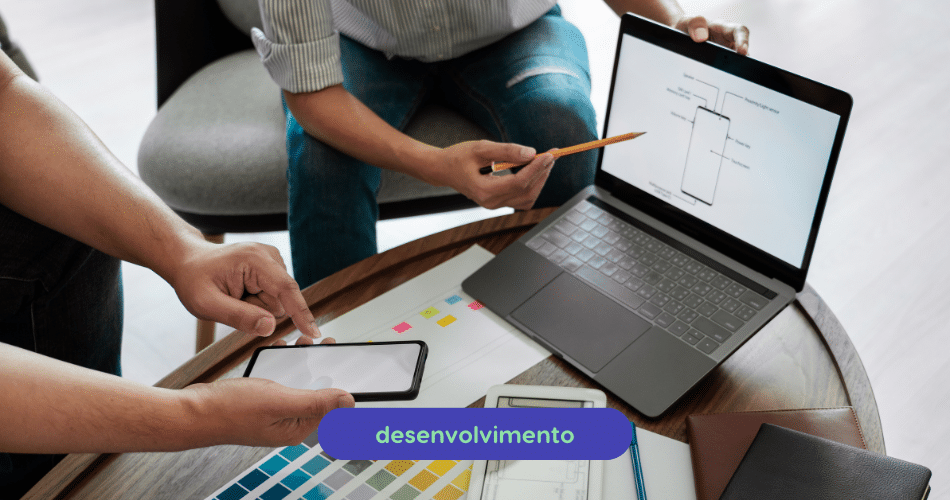 Duas pessoas sentam-se em uma mesa com um laptop exibindo um esquema de smartphone. Uma segura um telefone, enquanto outra aponta para a tela com um lápis. Uma paleta de cores e cadernos estão na mesa, focando no processo de "desenvolvimento MVP" na parte inferior.